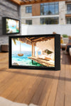 Vertical view of a weatherproof outdoor TV enclosure installed on a patio deck, showing high-definition tropical beach content on screen.